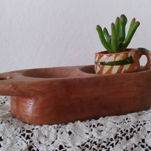 May include: A wooden serving dish with two compartments, one of which holds a small, orange and green ceramic pot with a succulent plant. The dish is sitting on a white lace doily.