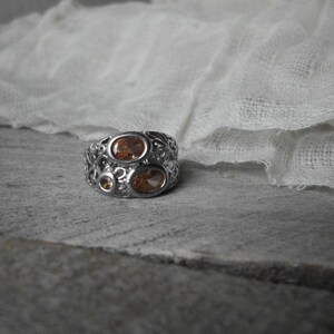 K&ouml;nnte beinhalten: Ein silberner Ring mit einem filigranen Design, das drei ovale orangefarbene Edelsteine ​​aufweist.