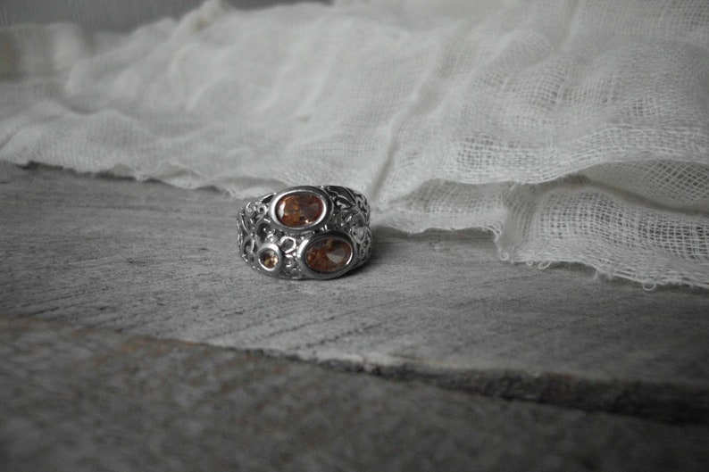 K&ouml;nnte beinhalten: Ein silberner Ring mit einem filigranen Design, das drei ovale orangefarbene Edelsteine ​​aufweist.