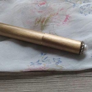 Puede incluir: Un bolígrafo o lápiz óptico cilíndrico de color dorado descansa sobre una tela blanca con estampado floral. El bolígrafo tiene un extremo decorativo con una punta de perla y un anillo de pequeñas piedras transparentes. El fondo es una superficie de madera clara.