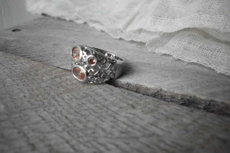 K&ouml;nnte beinhalten: Ein silberner Ring mit einem filigranen Design, das mit drei orangefarbenen Edelsteinen verziert ist.