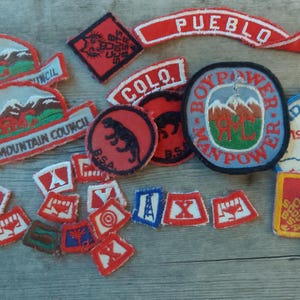 May include: A collection of vintage Boy Scouts of America patches. The patches feature various designs, including mountain landscapes, a panther, and the text "PUEBLO" and "ROCKY MOUNTAIN COUNCIL". The patches are in red, white, blue, and green.