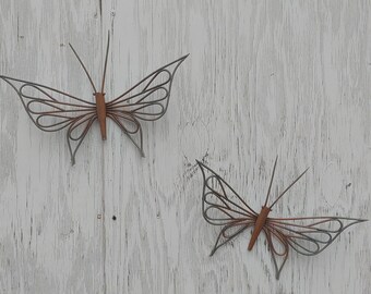 May include: Two metal butterflies with a rustic brown finish are attached to a white wooden wall. The butterflies are made of wire and have a delicate, intricate design.
