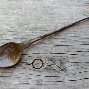 Puede incluir: Una cuchara rústica forjada a mano con un mango largo y delgado y un cuenco redondeado. La cuchara está hecha de metal y tiene una pátina marrón oscura.