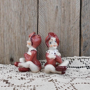 May include: Two ceramic figurines of people with red and white clothing sitting on a white crocheted surface. The figurines have red hats and white faces with black eyes and red lips.