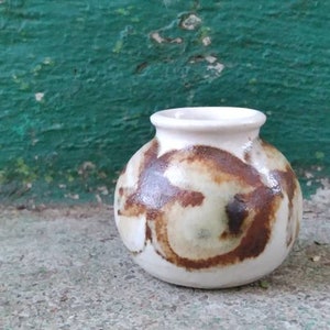 May include: A small, white ceramic vase with brown and white abstract designs. The vase is round and has a slightly flared opening.