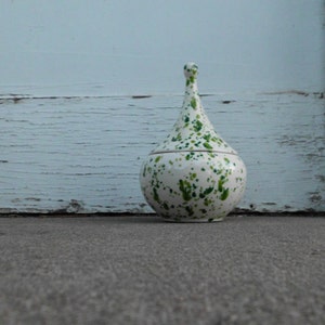 Op de afbeelding: Een witte keramische pot met een groen gespikkeld ontwerp. De pot heeft een rond lichaam en een lange, smalle hals.