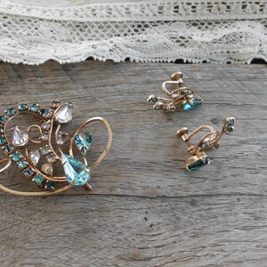 Op de afbeelding: Een vintage sieradenset met een broche en een paar oorclips. De set is gemaakt van goudkleurig metaal en heeft heldere en blauwe strass-steentjes in een bloemmotief.