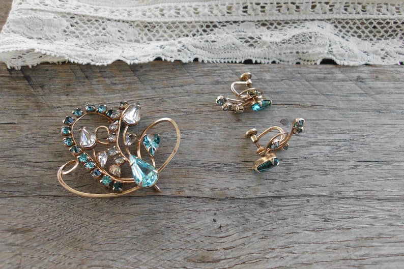 Op de afbeelding: Een vintage sieradenset met een broche en een paar oorclips. De set is gemaakt van goudkleurig metaal en heeft heldere en blauwe strass-steentjes in een bloemmotief.