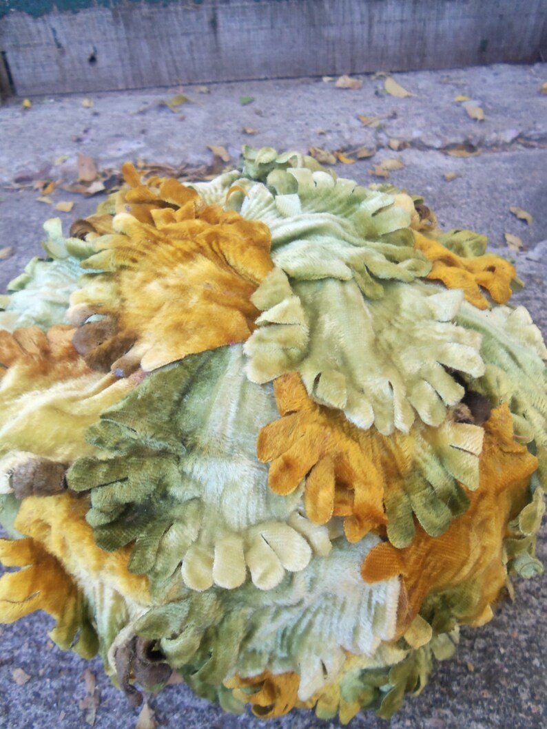 May include: A round, decorative object made of fabric with a textured, leafy design. The fabric is a mix of green, yellow, and brown colors.