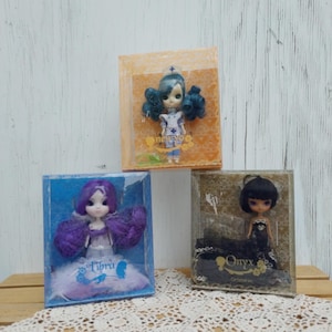May include: Three small dolls in display boxes. One doll has blue hair and wears a nurse's uniform. Another doll has purple hair and a dress with the word "Libra". The third doll has black hair and a black dress with the word "Onyx".
