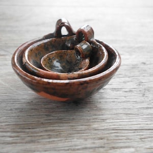 May include: A set of three nesting bowls made of brown and black speckled pottery. The bowls are glazed and have a rustic, handmade look.