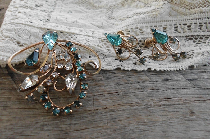 Op de afbeelding: Een vintage broche- en oorsteensets met een delicaat ontwerp van goudkleurig metaal met een cluster van heldere en tealblauwe strass.