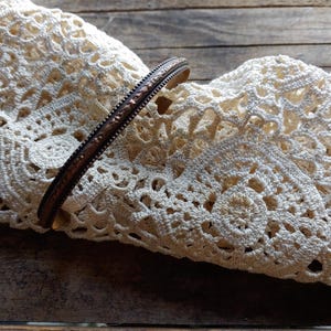 May include: A copper and brass bangle bracelet with a textured design. The bracelet is on a white crocheted doily.