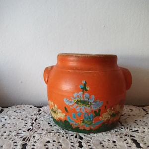 Puede incluir: Una olla de cerámica naranja con dos asas pequeñas y un diseño floral. Las flores están pintadas en azul, amarillo y verde. La olla está sobre un mantel individual de encaje blanco.