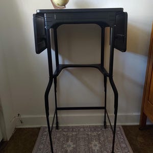 May include: A black metal side table with drop-leaf sides and a lower shelf, resting on small wheels. A decorative globe sits on the tabletop. The table is positioned on a patterned rug, creating a functional and stylish accent piece.