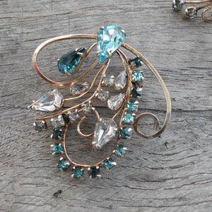 Op de afbeelding: Een paar gouden broche's met een delicaat, wervelend ontwerp. De broche's zijn versierd met heldere en turquoise blauwe strass.