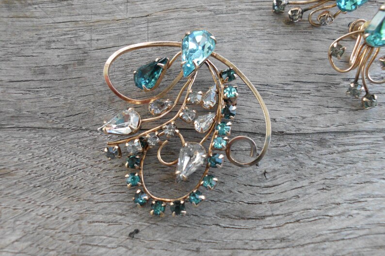 Op de afbeelding: Een paar gouden broche's met een delicaat, wervelend ontwerp. De broche's zijn versierd met heldere en turquoise blauwe strass.