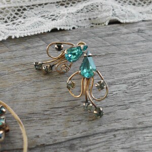 Op de afbeelding: Een paar gouden oorbellen met druppelvormige turquoise blauwe strass steentjes. De oorbellen hebben een delicaat, ingewikkeld ontwerp.