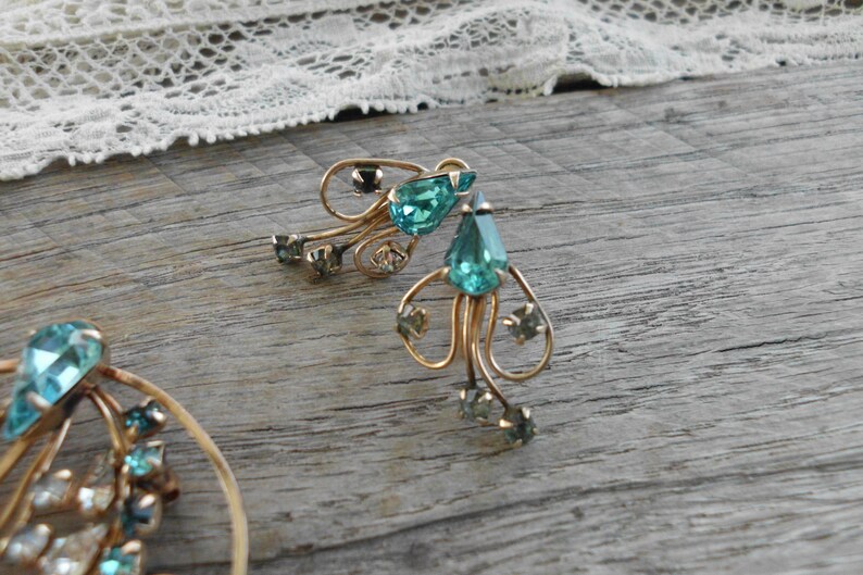 Op de afbeelding: Een paar gouden oorbellen met druppelvormige turquoise blauwe strass steentjes. De oorbellen hebben een delicaat, ingewikkeld ontwerp.