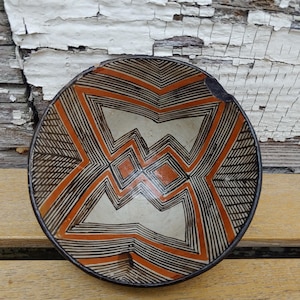 May include: A small, round ceramic bowl featuring a geometric pattern of black lines and orange shapes on a cream-colored background. The interior design consists of symmetrical, angular motifs radiating from the center, with a small chip on the rim.