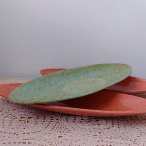May include: Two ceramic serving dishes, one green and one orange, are stacked on top of each other. The dishes are oval shaped and have a speckled glaze.