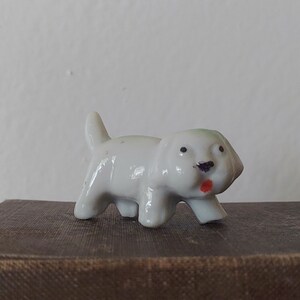 May include: A small white porcelain figurine of a dog with a red tongue. The dog is walking with its head tilted to the side.