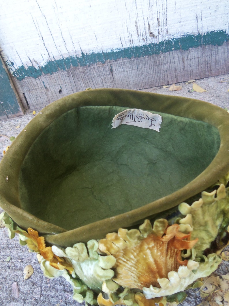 May include: A green velvet hat with a label that reads "Jacqueline II, Designed by Sally Victor". The hat is decorated with velvet leaves in shades of green, yellow, and orange.