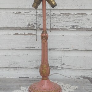 Peut inclure: Une lampe de table vintage rose avec des accents dorés et trois douilles. La lampe a une base décorative et une tige fine.