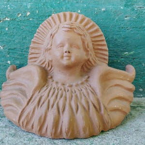 May include: A terracotta-colored wall plaque featuring a sculpted cherub head with a radiating halo design behind it. The cherub has detailed facial features and is framed by stylized, sweeping wings that extend outward on both sides.