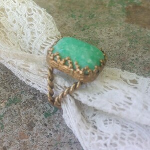May include: A gold ring with a green gemstone set in a decorative frame. The ring is on a white lace background.