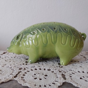 May include: A light green ceramic hedgehog ornament with decorative swirls and holes on its back. The figurine has a glossy finish and is on a white lace doily.