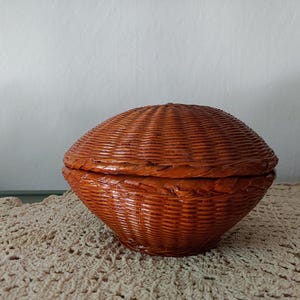 May include: A round, lidded basket made of woven brown material. The basket has a textured surface and a domed lid. The basket is sitting on a cream-colored, crocheted doily.