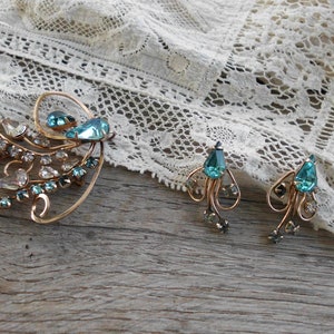Op de afbeelding: Een vintage sieradenset met een grote broche en een paar oorbellen. De set is gemaakt van goudkleurig metaal en is bezet met blauwe en heldere strasssteentjes. De broche heeft de vorm van een gestileerde bloem met een grote blauwe strasssteen in het midden. De oorbellen zijn hangende oorbellen met een blauwe strasssteen-druppel.
