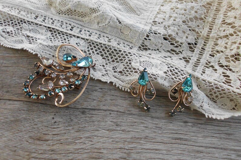 Op de afbeelding: Een vintage sieradenset met een grote broche en een paar oorbellen. De set is gemaakt van goudkleurig metaal en is bezet met blauwe en heldere strasssteentjes. De broche heeft de vorm van een gestileerde bloem met een grote blauwe strasssteen in het midden. De oorbellen zijn hangende oorbellen met een blauwe strasssteen-druppel.