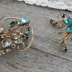 Op de afbeelding: Een paar vintage gouden oorbellen met een delicaat, wervelend ontwerp. De oorbellen zijn versierd met heldere en teal blauwe strass.