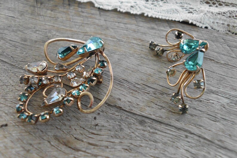 Op de afbeelding: Een paar vintage gouden oorbellen met een delicaat, wervelend ontwerp. De oorbellen zijn versierd met heldere en teal blauwe strass.