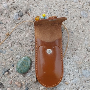 May include: A tan leather pouch with a press stud closure, open to show the inside. A small, polished green stone is next to it. The pouch has a beaded charm in yellow, orange, and blue.