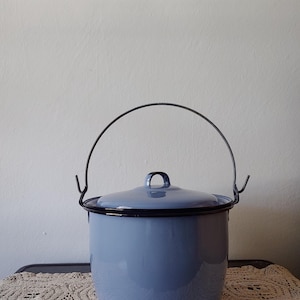Może przedstawiać: Jasnoniebieski emaliowany garnek z pokrywką i czarnym drucianym uchwytem. Garnek ma zaokrąglony kształt i czarną obwódkę. Pokrywka ma mały uchwyt w kształcie pętli. Garnek stoi na stole z koronkowym obrusem.