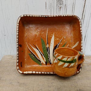May include: Hand-painted, square terracotta tray and matching mug. The tray features a design of agave plants in green and white against an orange background. The mug has green and white stripes. The set is on a wooden surface.