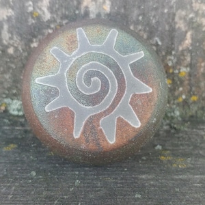 May include: A round, ceramic sun with a spiral design in the centre. The sun is painted in shades of brown, green, and white.