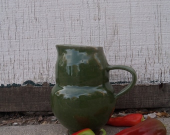 May include: A green ceramic pitcher with a wide mouth and a handle. The pitcher is sitting on a concrete surface with three red peppers.