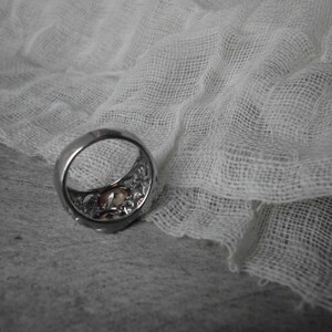 K&ouml;nnte beinhalten: Ein silberner Ring mit einem dekorativen Design und einem klaren Stein in der Mitte. Der Ring liegt auf einem wei&szlig;en Stoffhintergrund.