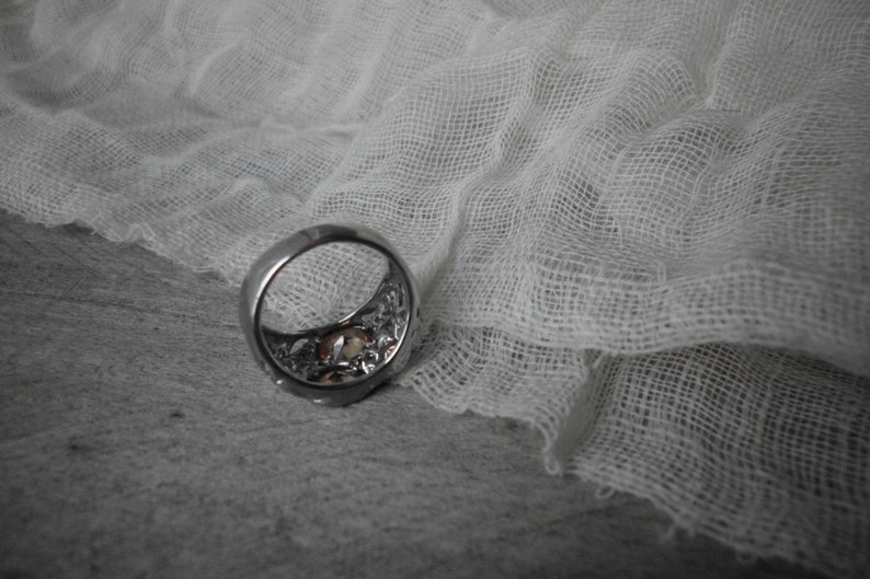 K&ouml;nnte beinhalten: Ein silberner Ring mit einem dekorativen Design und einem klaren Stein in der Mitte. Der Ring liegt auf einem wei&szlig;en Stoffhintergrund.