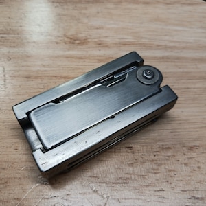May include: A silver multi-tool with a brushed metal finish. The tool is closed and has a small round button on the side.