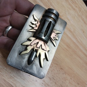 May include: A silver belt buckle with a stylized design of a bullet with a copper and gold flame behind it.