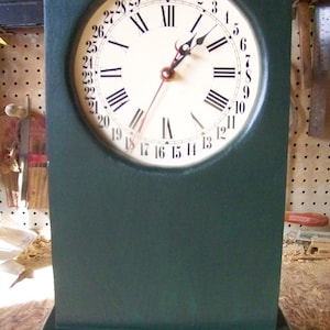 May include: A dark green wooden clock with a white face and black Roman numeral hour markers. The clock face also has black numbers for the days of the month, with the numbers 20 through 31 visible. The clock hands are black with a red second hand.