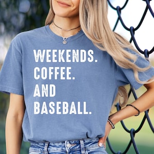 May include: A blue t-shirt with the words "WEEKENDS. COFFEE. AND BASEBALL." in white block letters. The shirt is short-sleeved and has a slightly faded appearance. The person is wearing a necklace and bracelets.