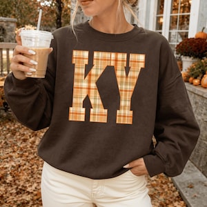May include: A dark brown long-sleeve crewneck sweatshirt featuring the letters KY in a large block font. The letters are filled with an orange, yellow, and white plaid pattern. The sweatshirt is paired with light-coloured trousers.