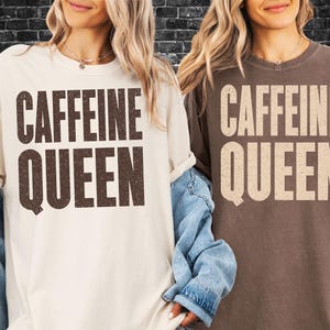 May include: Two t-shirts, one cream-colored and one brown, each with the words "CAFFEINE QUEEN" printed in a bold, distressed font. The shirts are casual and have short sleeves.